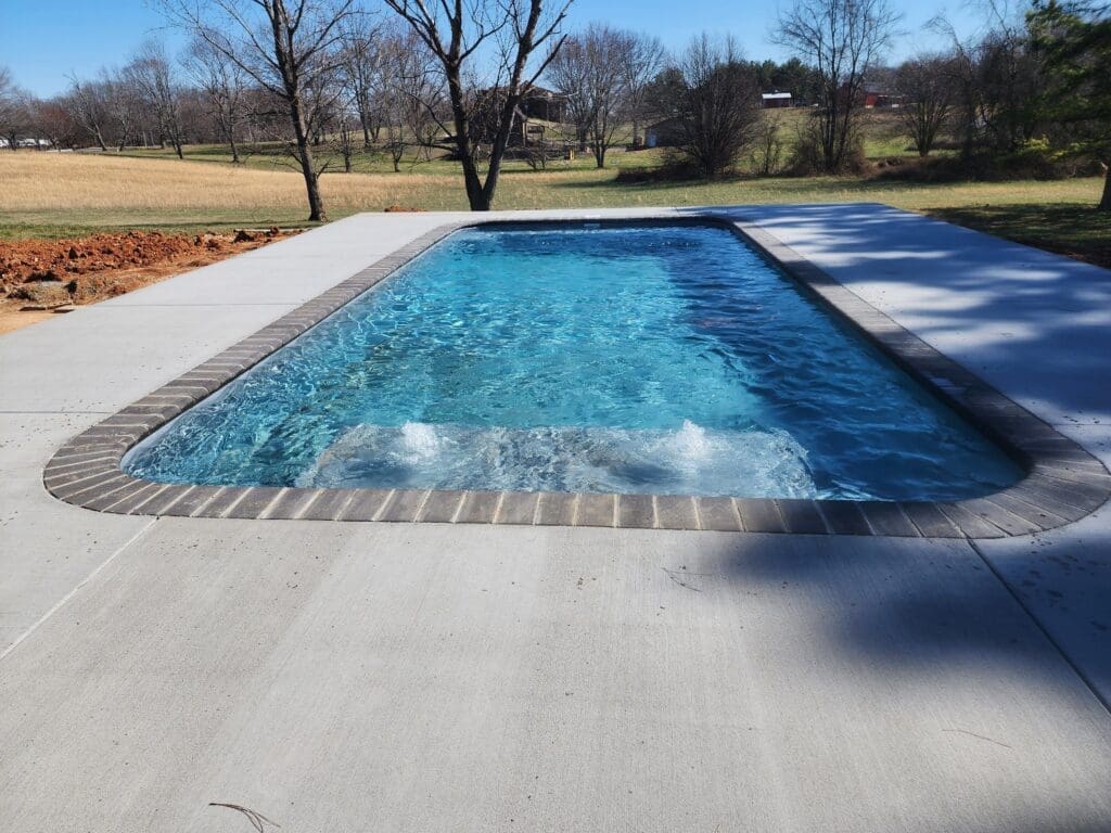 Rectangular inground pool with clear blue water on a sunny day with grass and trees in the background.