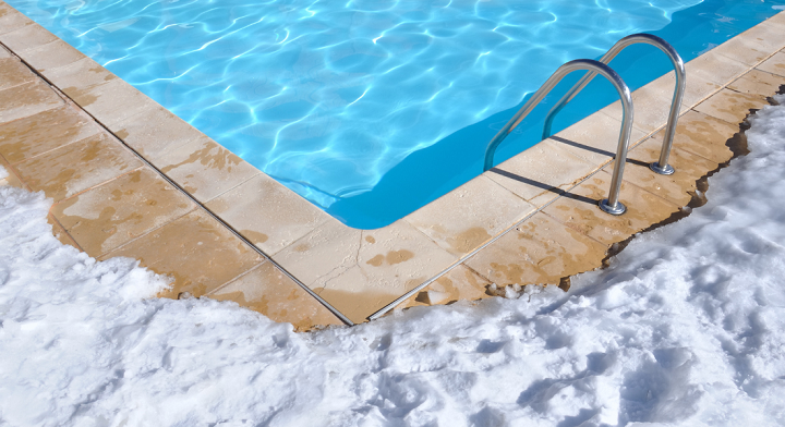 A swimming pool surrounded by snow, with a metal ladder leading into the water, awaits closing for winter.