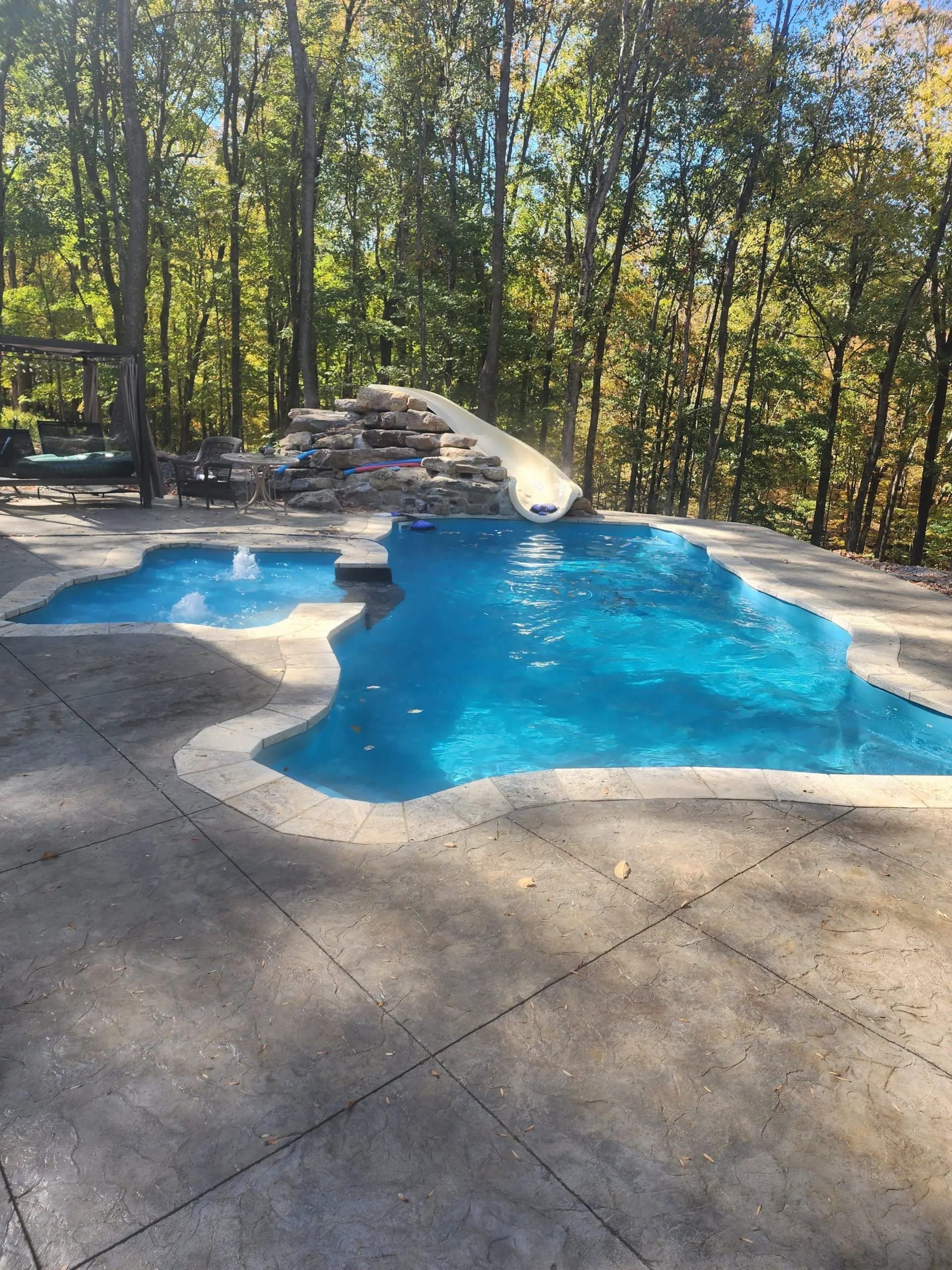 A backyard swimming pool with a slide and surrounding patio area amidst trees displaying autumn foliage.