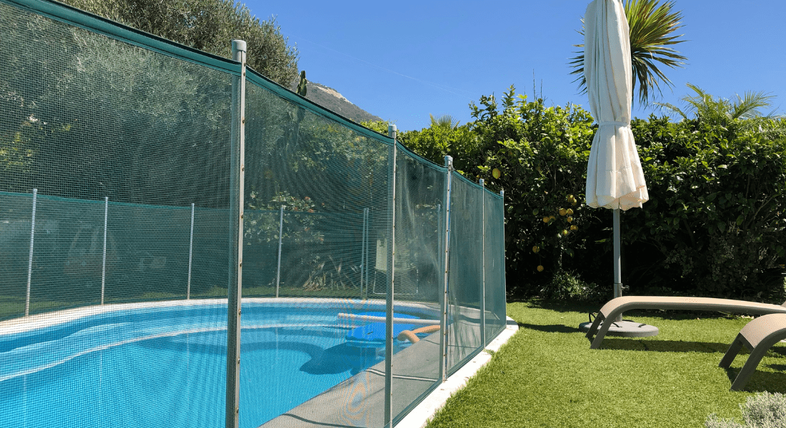 A garden featuring a sunlit oval swimming pool enclosed by a safety fence with a white umbrella and lounge chairs on a grassy area.