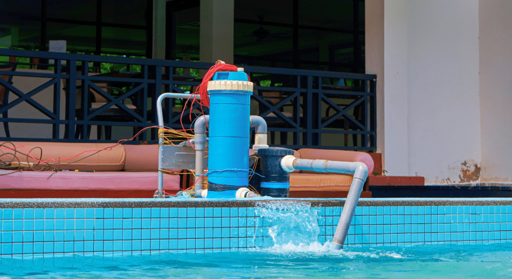 A blue pool filtration system with connected pipes and wires is set up at the edge of a swimming pool, next to a building with chairs and a railing. Water is being filtered and returned to the pool.