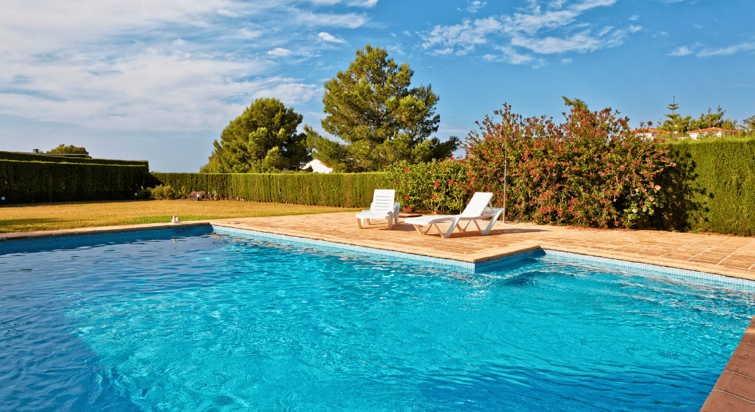 A clear blue swimming pool with two white lounge chairs on a brick-paved deck. The area is surrounded by greenery, including hedges and trees, under a partially cloudy sky.