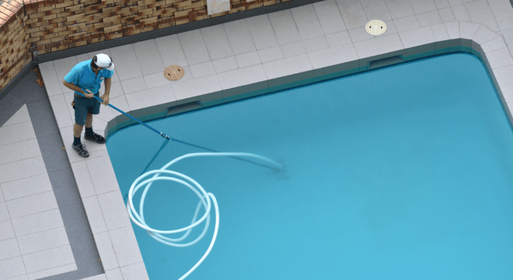 A person wearing a blue shirt and hat is cleaning a swimming pool with a long pole vacuum cleaner. The area around the pool has tiled flooring. Pool Cleaning Services 