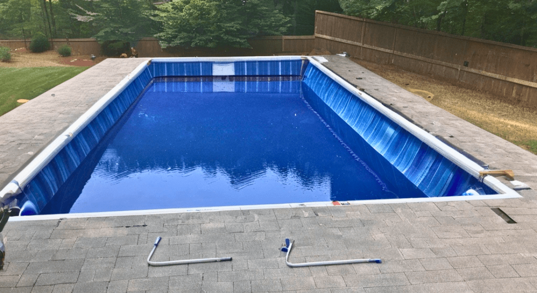 A rectangular inground swimming pool surrounded by a paved area with greenery and a wooden fence in the background. Two pool cleaning tools are placed on the pavers near the pool. pool liner replacement