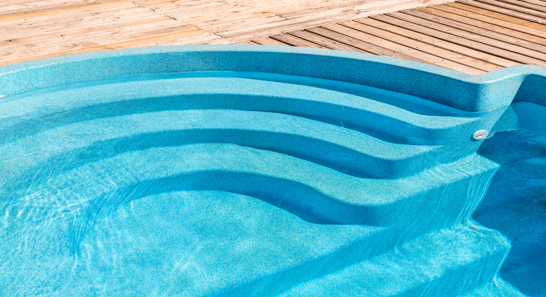 Blue swimming pool with curved steps and a wooden deck in the background. Custom Fiberglass pool