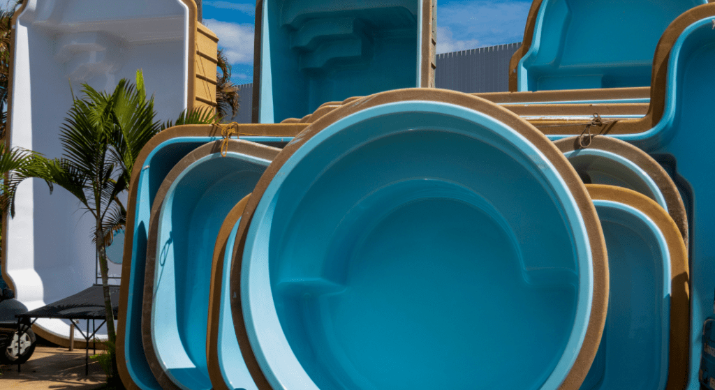 A collection of stacked, empty fiberglass swimming pool shells in various shapes and sizes, displayed outdoors under a clear blue sky. Custom Fiberglass Pool