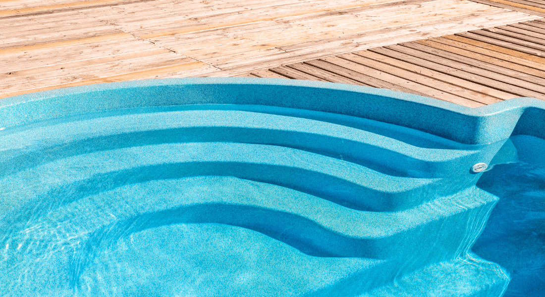 Close-up of a turquoise swimming pool with curved steps and a wooden deck. how much fiberglass pool cost.