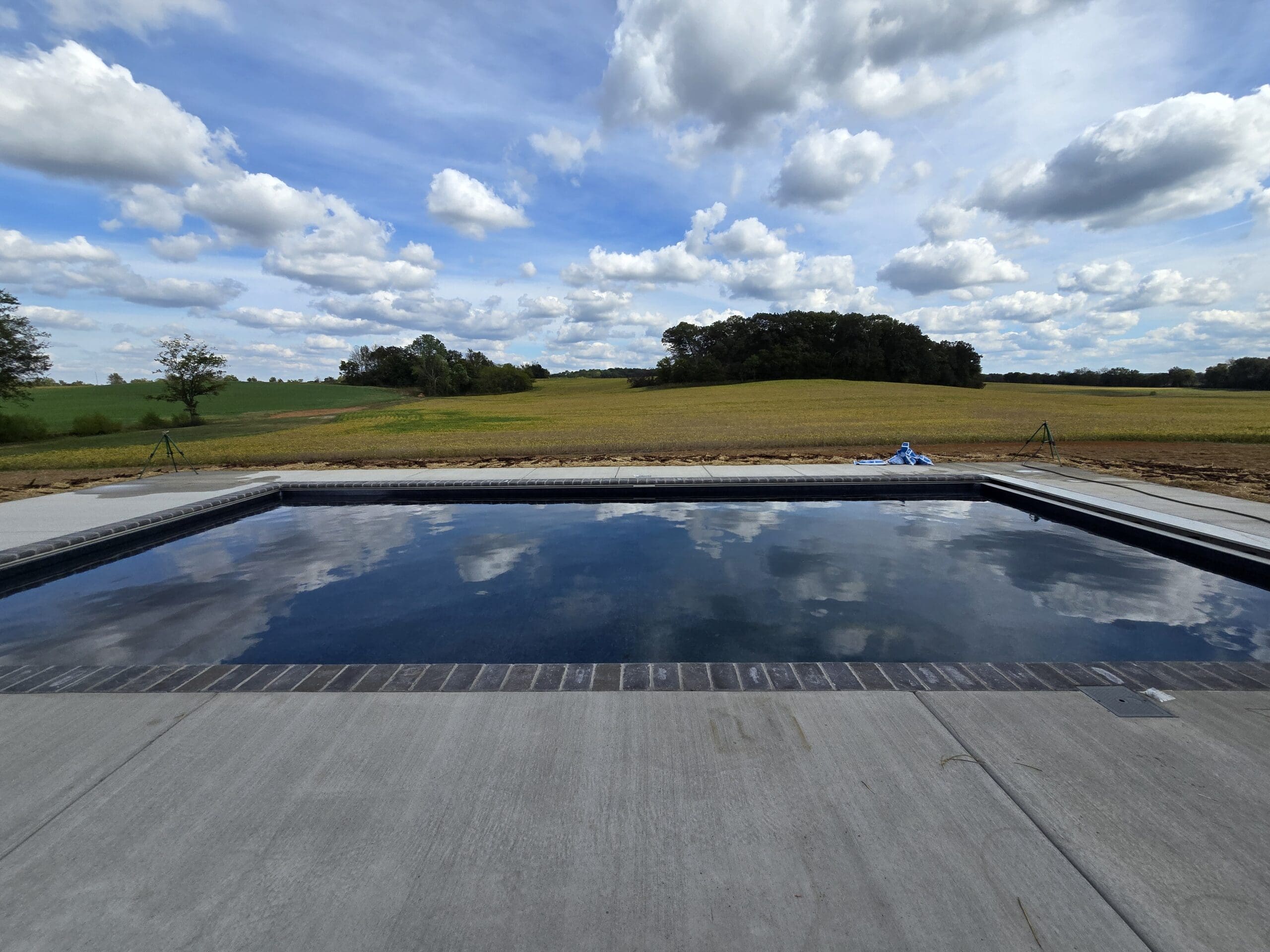 Rectangular outdoor swimming pool with calm water reflecting a partly cloudy sky, surrounded by concrete deck and open fields in the background.
