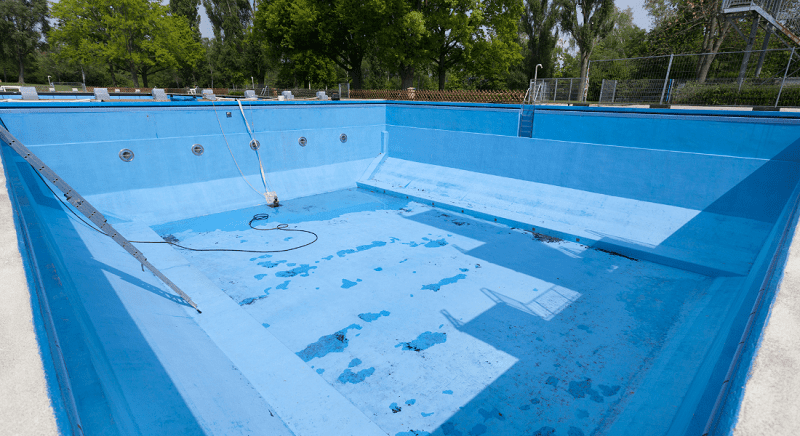 An empty outdoor swimming pool with peeling blue paint and a small puddle of water at the bottom. A metal ladder and some maintenance equipment are visible.