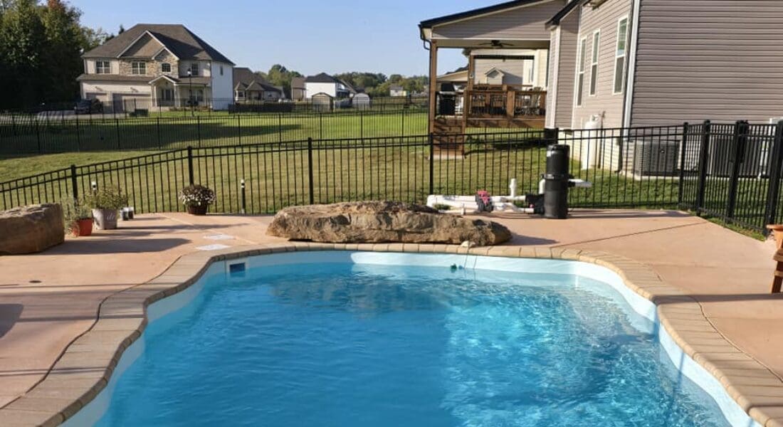A backyard swimming pool with a stone border, surrounded by a black metal fence, with houses and a grassy yard in the background.