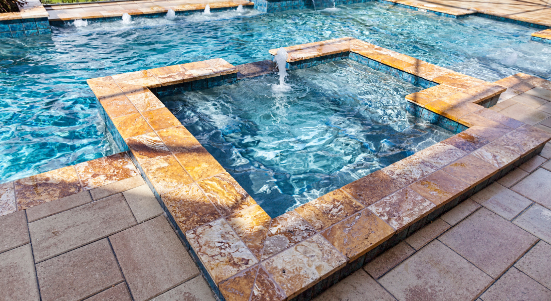 A tiled spa with a small fountain sits adjacent to a swimming pool, both surrounded by tan stone pavers.