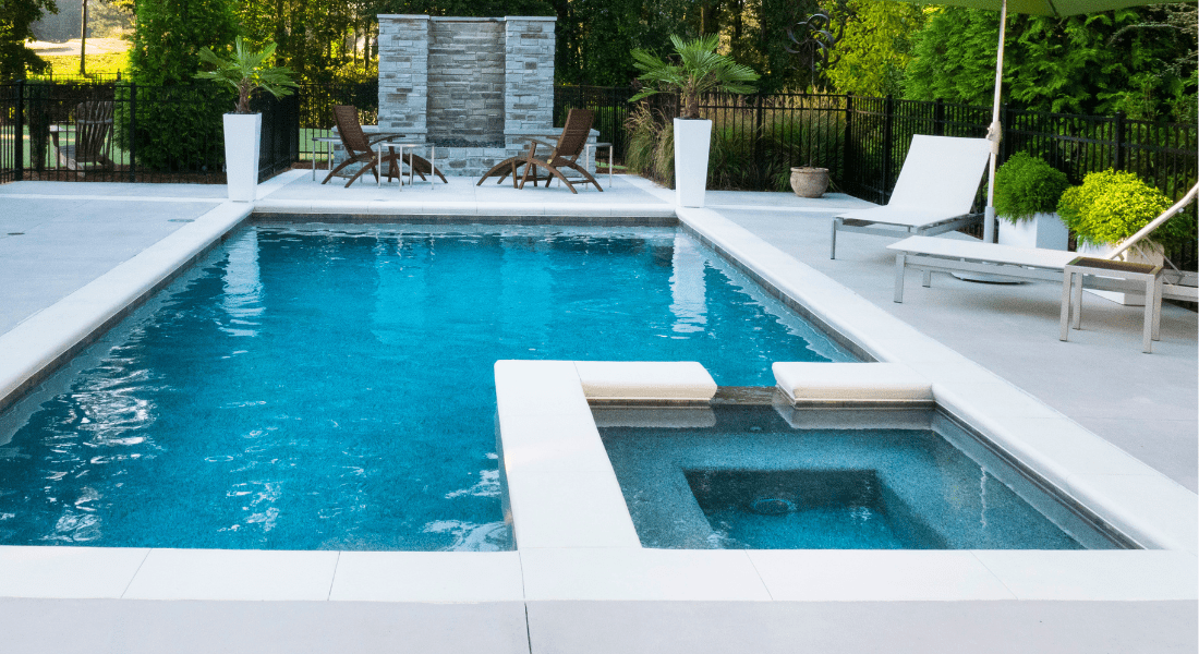 A modern outdoor swimming pool with an attached hot tub, lounge chairs, and a stone water feature, surrounded by concrete decking and greenery.