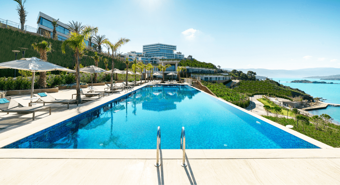Rectangular outdoor swimming pool with lounge chairs and umbrellas, surrounded by greenery, overlooking the sea and modern buildings in the background on a sunny day.