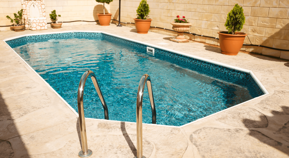Rectangular outdoor swimming pool with metal ladder, surrounded by potted plants and beige stone tiles.